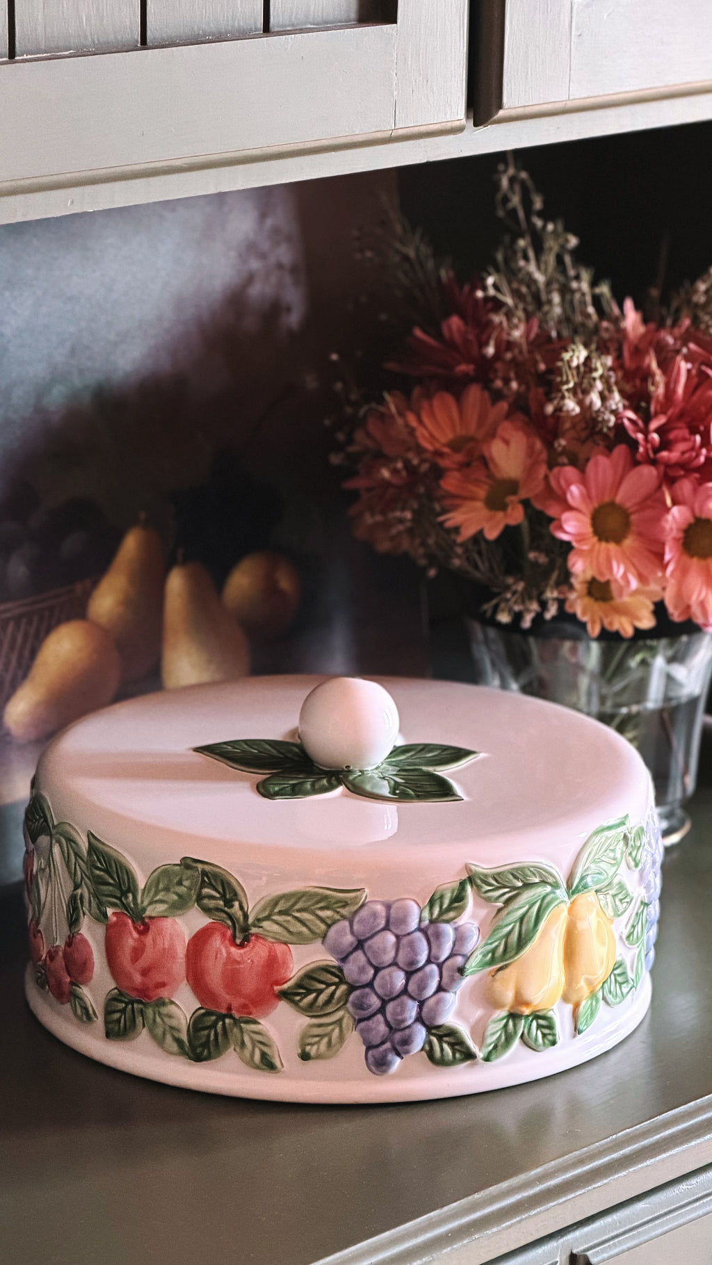 A round ceramic cake or cheese cover with a lid, decorated with raised 3D fruit motifs including red apples, purple grapes, yellow pears, and green leaves. The cover is shown on a kitchen counter with dried flowers and pears in the background.