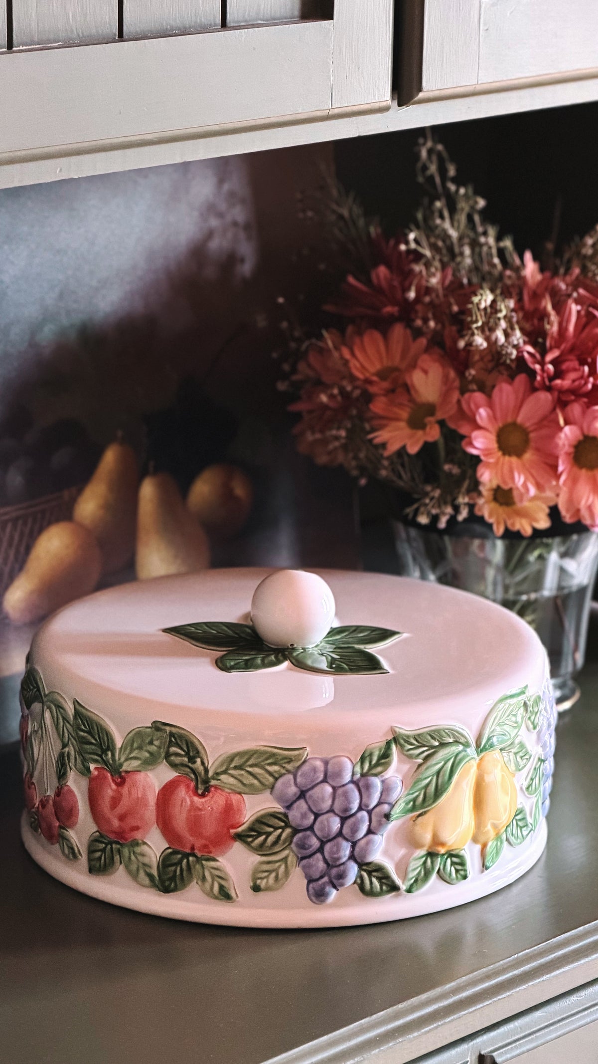 A round ceramic cake or cheese cover with a lid, decorated with raised 3D fruit motifs including red apples, purple grapes, yellow pears, and green leaves. The cover is shown on a kitchen counter with dried flowers and pears in the background.
