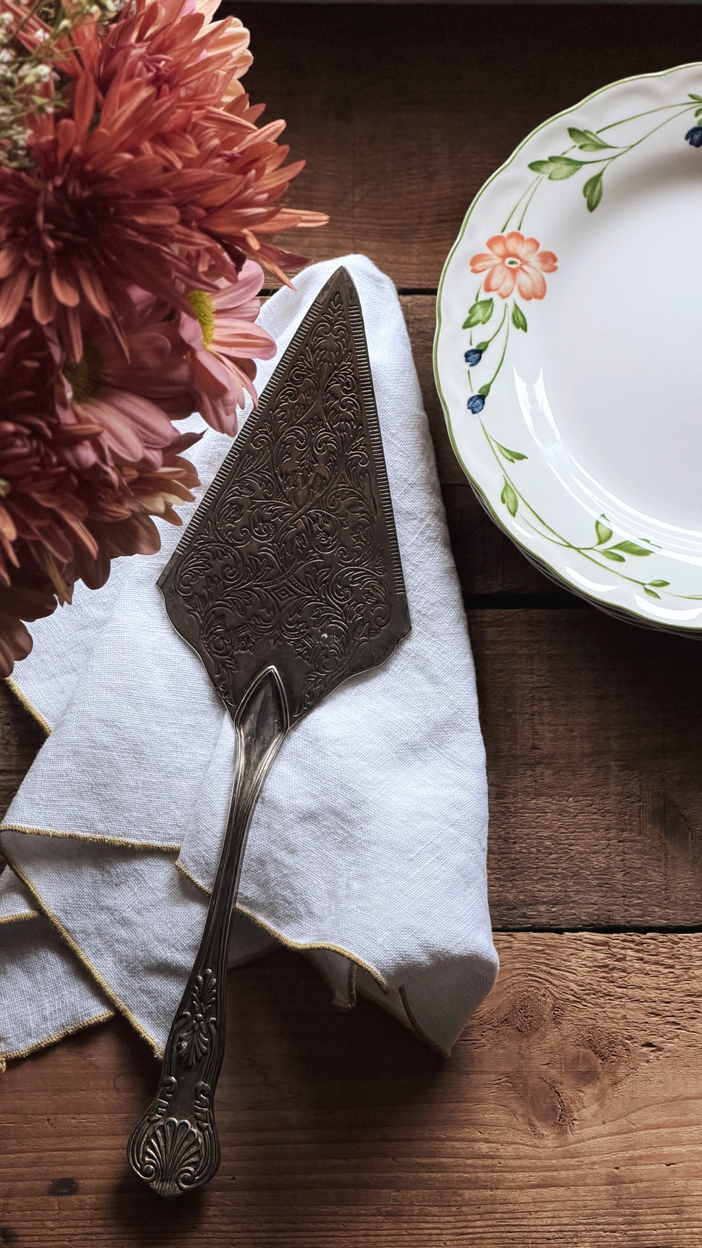 An ornate vintage-style metal cake or dessert server resting on a white napkin on a rustic wooden table, with pink flowers on the left and part of a floral ceramic plate on the right.
