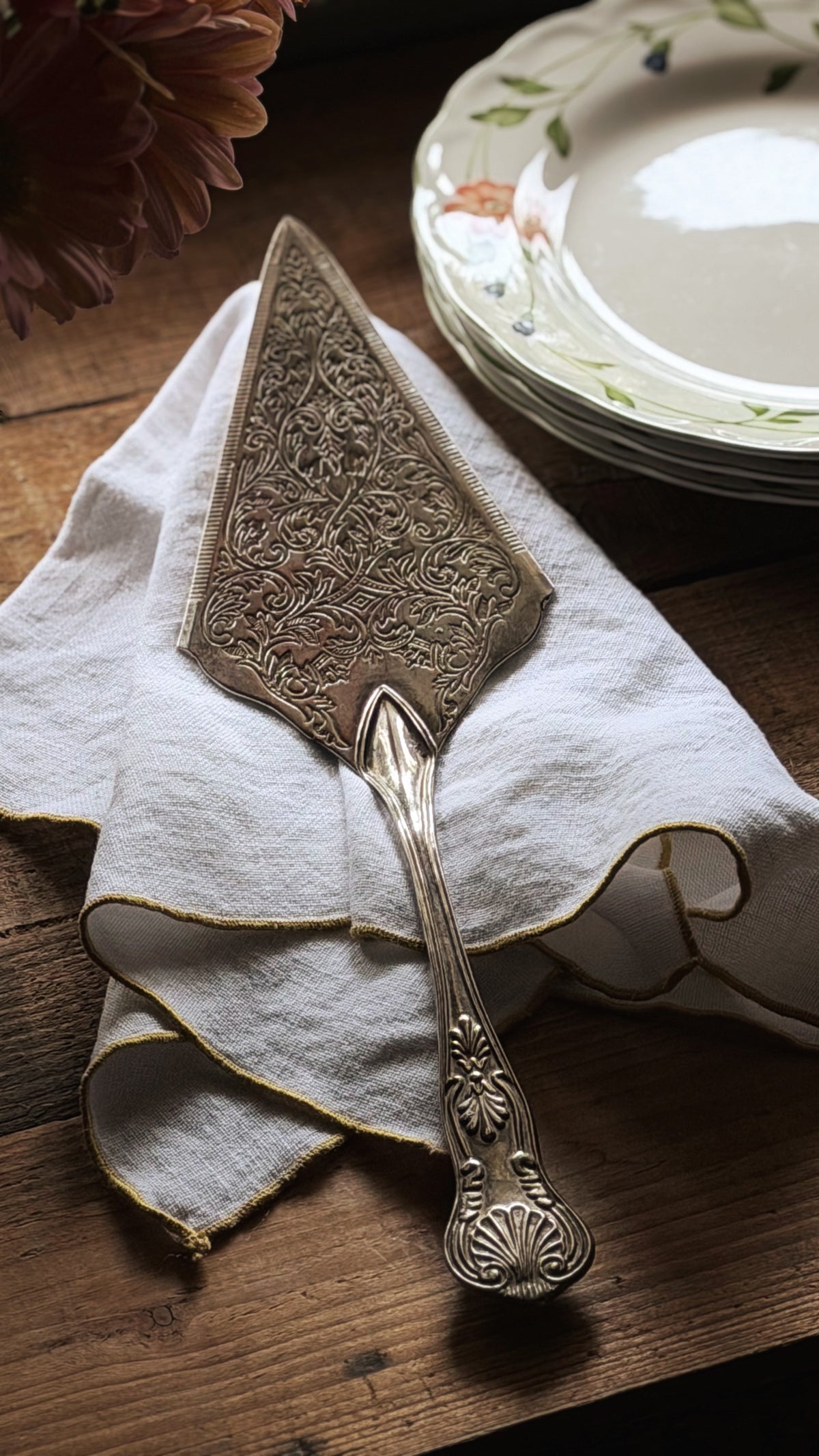 An ornate vintage silver cake pie server with intricate floral engraving, resting on a white linen napkin on a rustic wooden table, with stacked floral plates in the background.