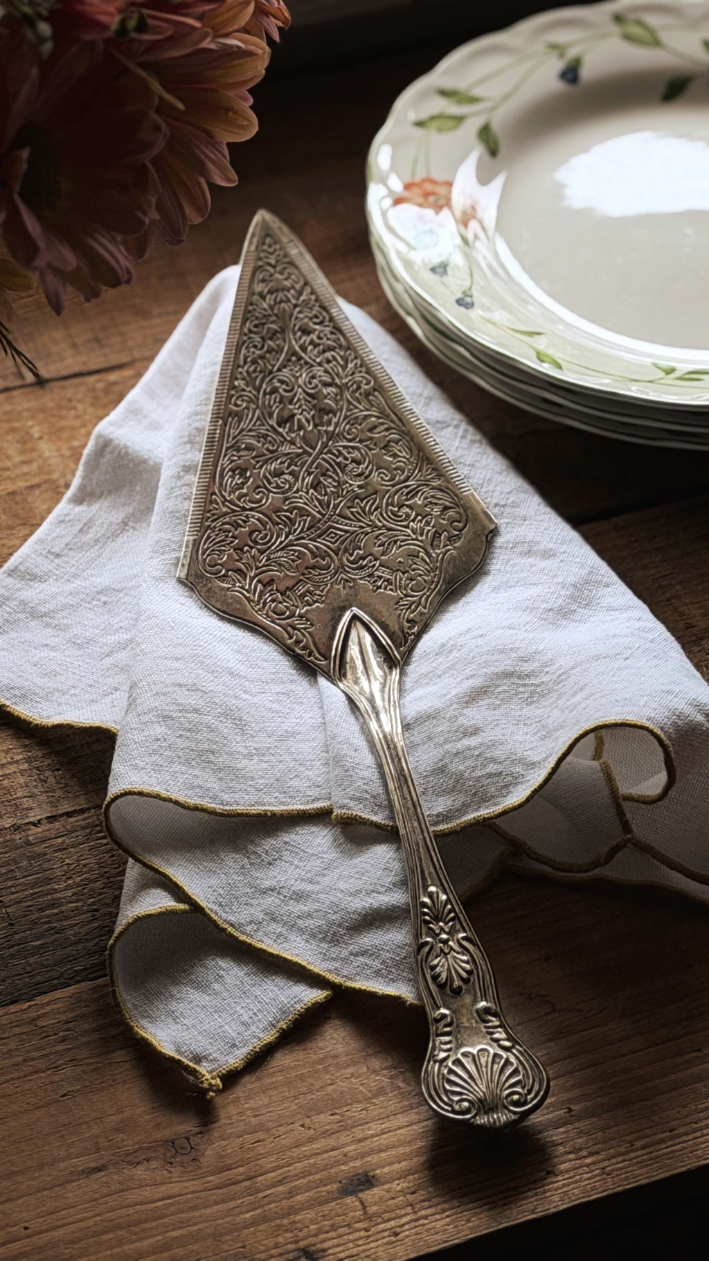 An ornate silver cake server with intricate floral engravings, resting on a white napkin with gold trim on a rustic wooden table, with stacked plates and flowers in the background.