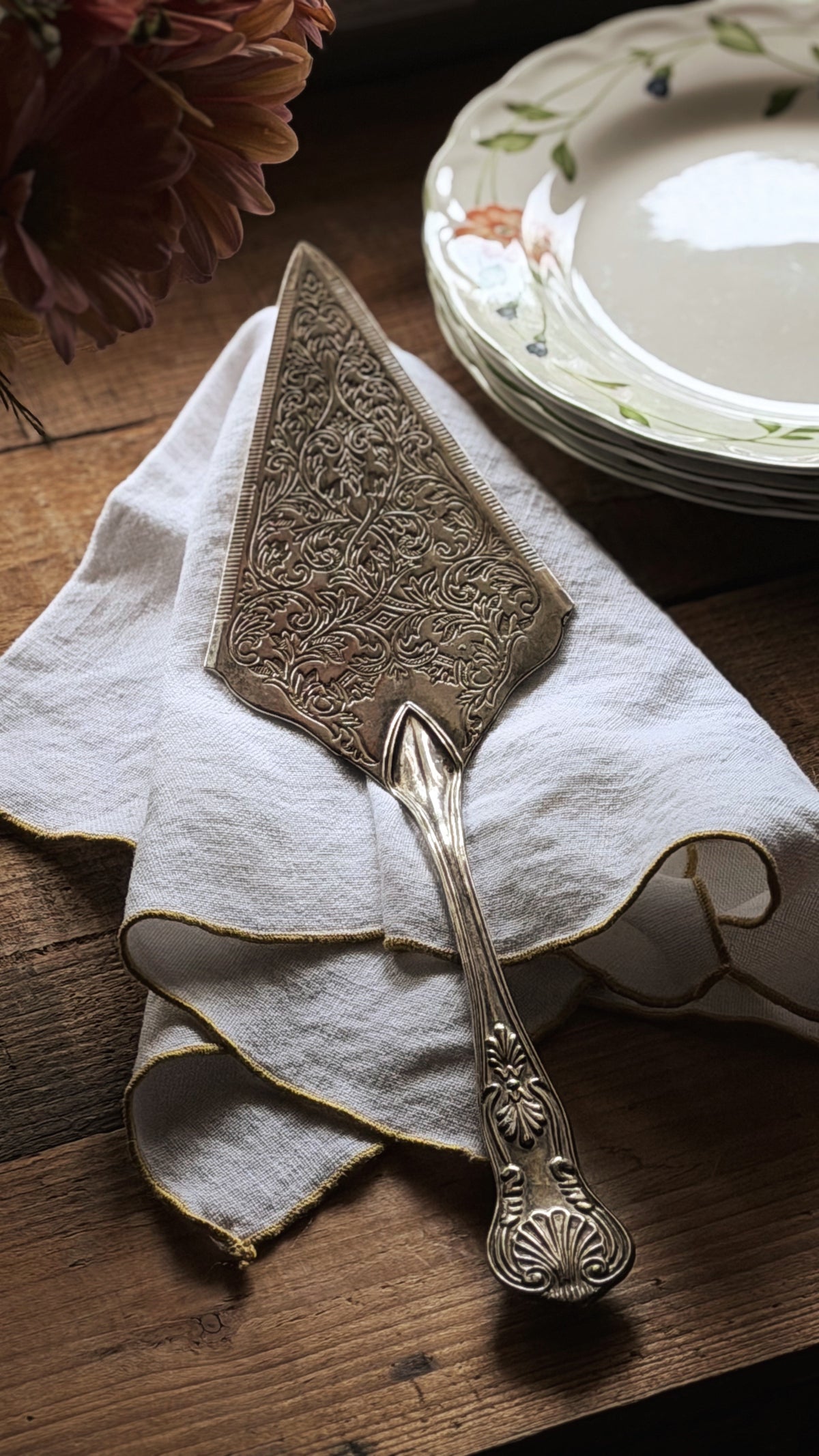 An ornate silver cake server with intricate floral engravings, resting on a white napkin with gold trim on a rustic wooden table, with stacked plates and flowers in the background.