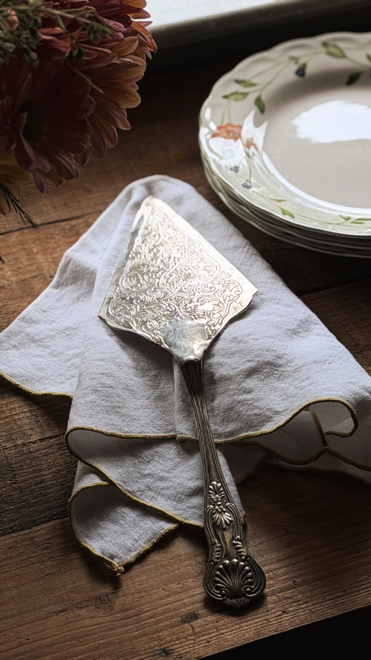 An ornate vintage silver cake or dessert server resting on a light beige linen napkin with gold trim, set on a rustic wooden table next to a stack of floral ceramic plates.
