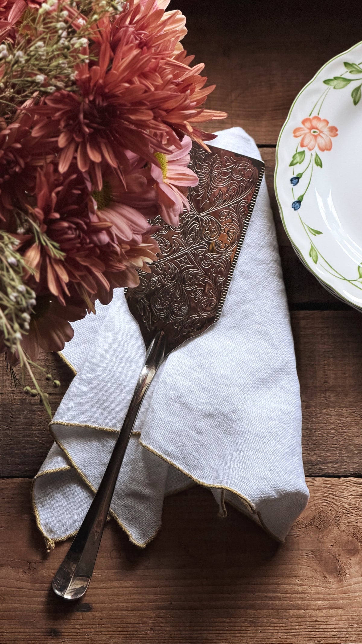 An ornate silver metal cake server with intricate embossed floral detailing, resting on a folded white linen napkin on a rustic wooden table, next to a bouquet of pink chrysanthemums and a floral ceramic plate.