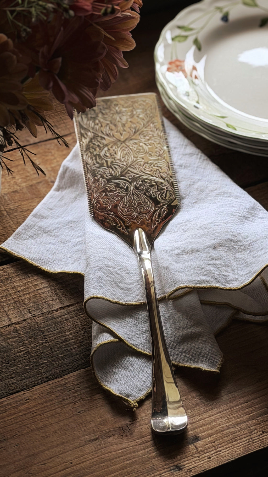 An ornate silver cake or cheese server with a detailed embossed floral pattern, resting on a white linen napkin on a rustic wooden table.