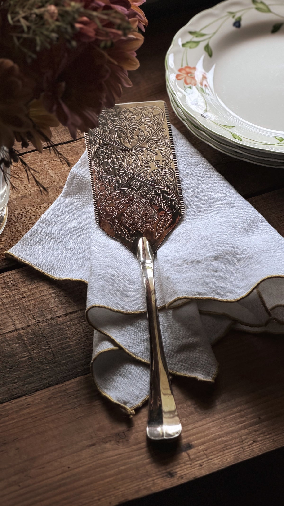 An ornate metal cake server with intricate embossed detailing, resting on a white linen napkin on a rustic wooden table, with a stack of floral plates in the background.