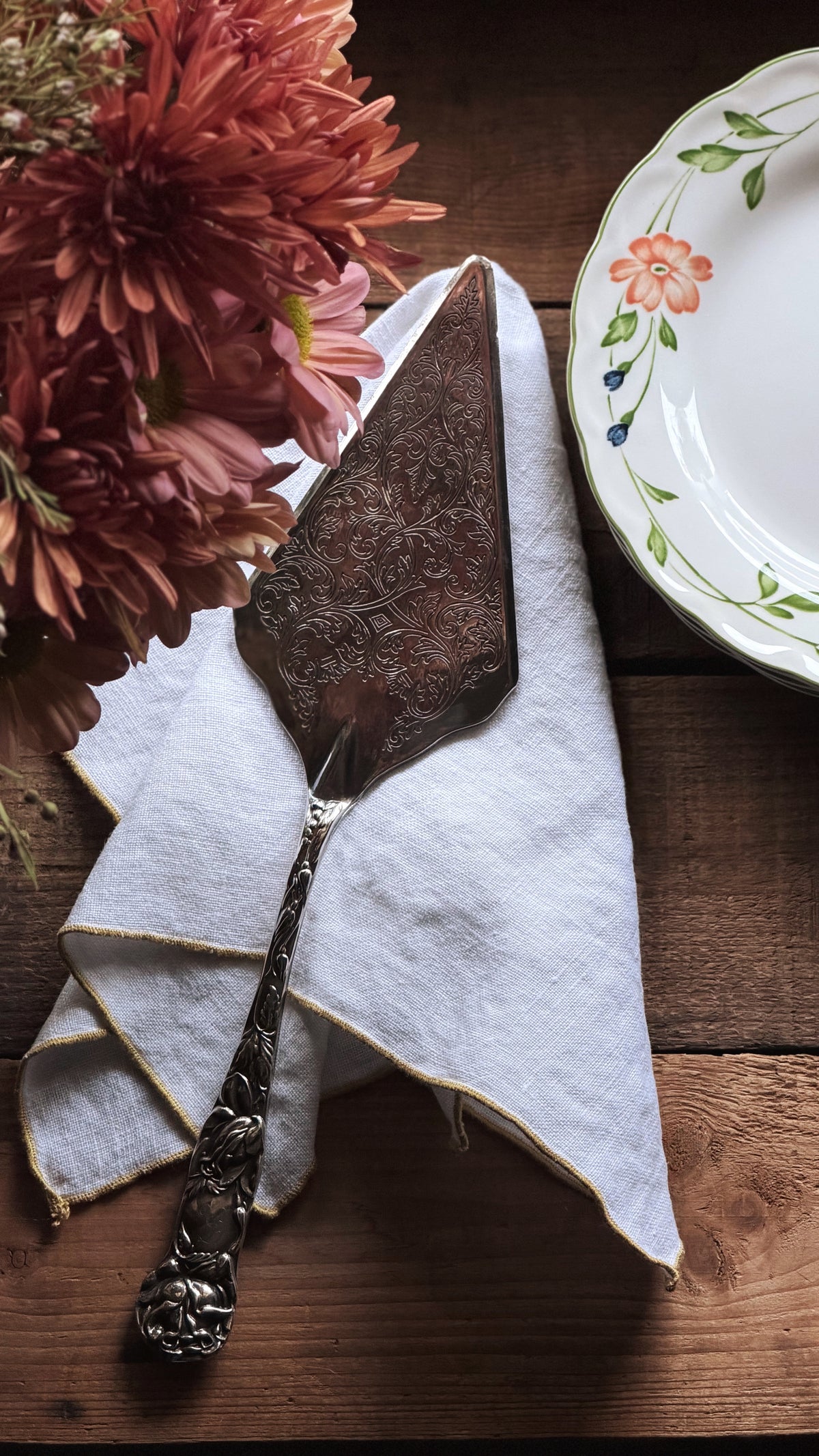 An ornate vintage-style metal cake server resting on a white napkin with gold trim, set on a rustic wooden table alongside a floral plate and pink flowers.