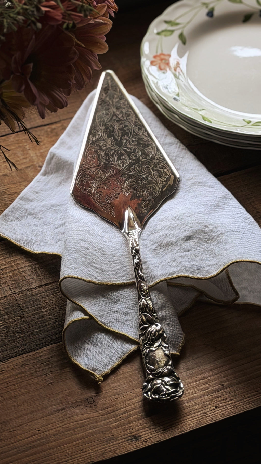 A vintage ornate silver cake or cheese server with a patterned blade, resting on a white linen napkin on a dark wooden table, with a stack of floral plates in the background.
