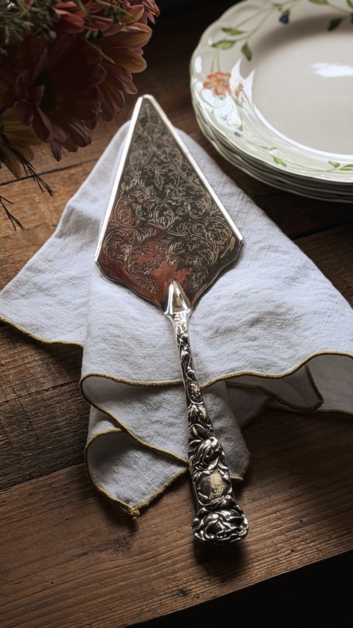 A vintage ornate silver cake or cheese server with a patterned blade, resting on a white linen napkin on a dark wooden table, with a stack of floral plates in the background.