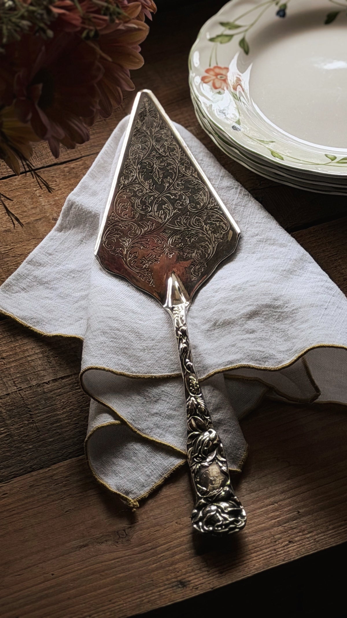 An ornate vintage silver cake or pie server with a highly detailed handle, resting on a white linen napkin on a dark wooden table. A stack of floral ceramic plates is visible in the background.