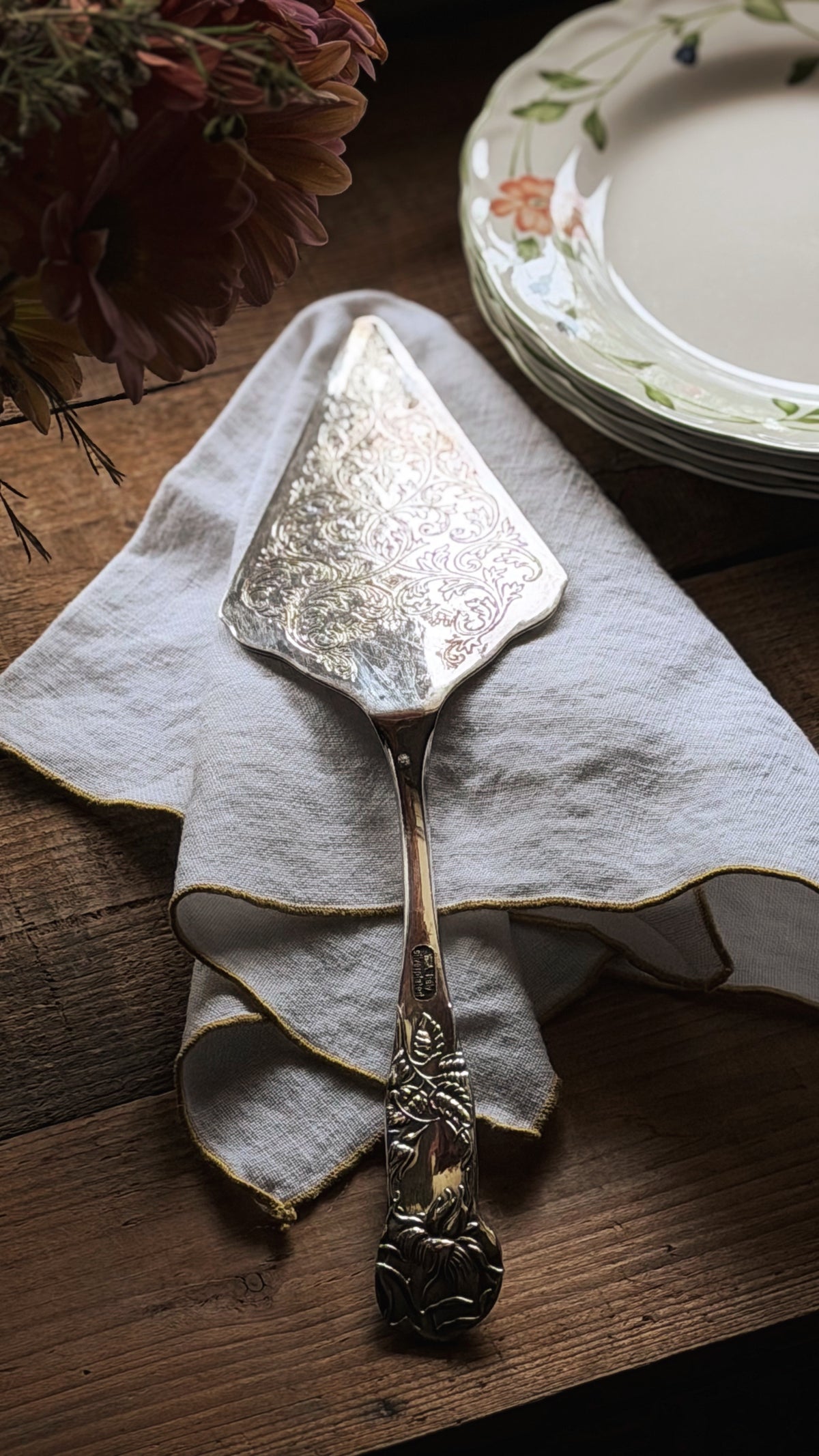 An ornate vintage silver cake server with a floral handle, resting on a folded cream linen napkin on a rustic wooden table, with plates and dried flowers in the background.