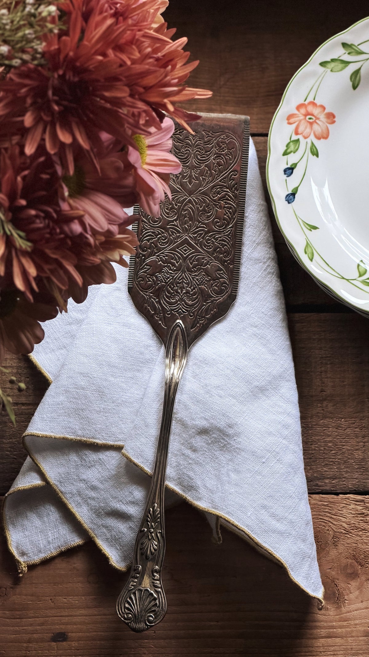 An ornate silver-toned metal cake server with embossed detailing rests on a folded white linen napkin on a dark wooden table, styled with a floral arrangement and part of a floral ceramic plate.