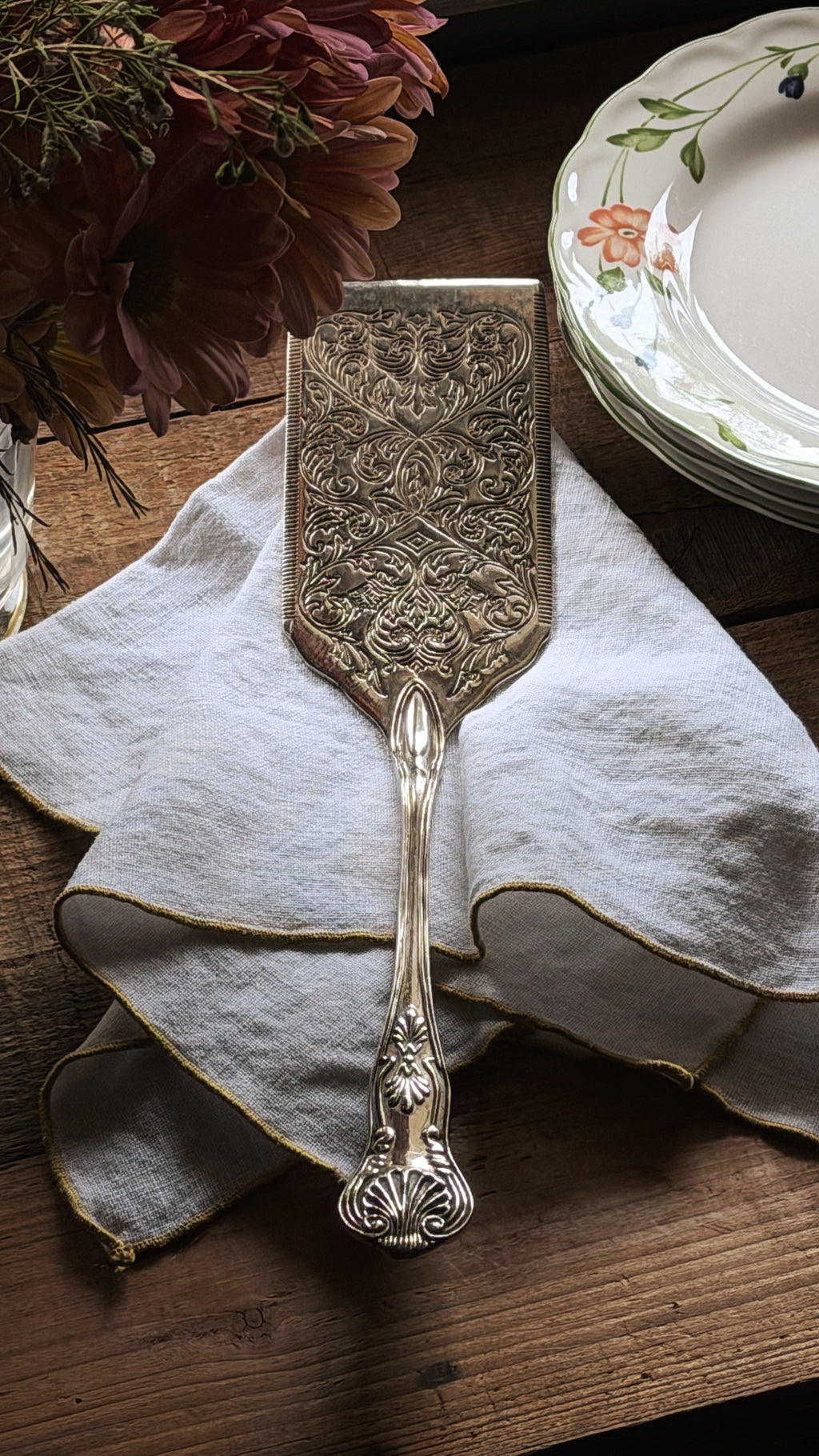An ornate silver-toned metal cake server with intricate embossed detailing, resting on a white linen napkin on a rustic wooden table, with a stack of floral plates and dried flowers in the background.