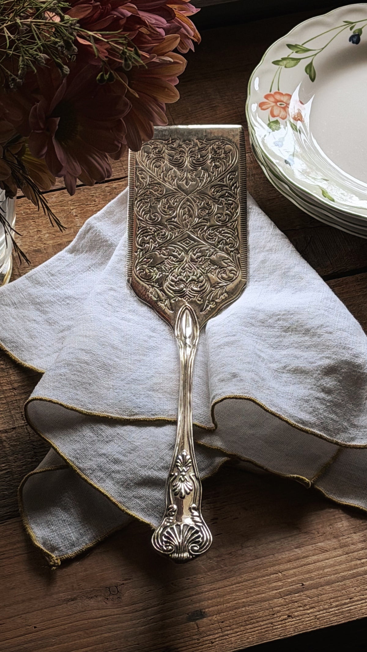 An ornate silver-toned metal cake server with intricate embossed detailing, resting on a white linen napkin on a rustic wooden table, with a stack of floral plates and dried flowers in the background.