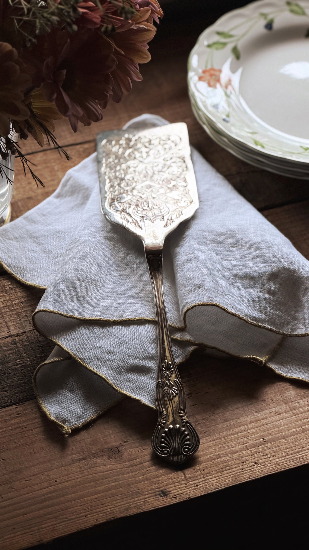 An ornate vintage-style metal cake server or serving spatula with an engraved floral pattern, resting on a folded cream linen napkin on a rustic wooden table.