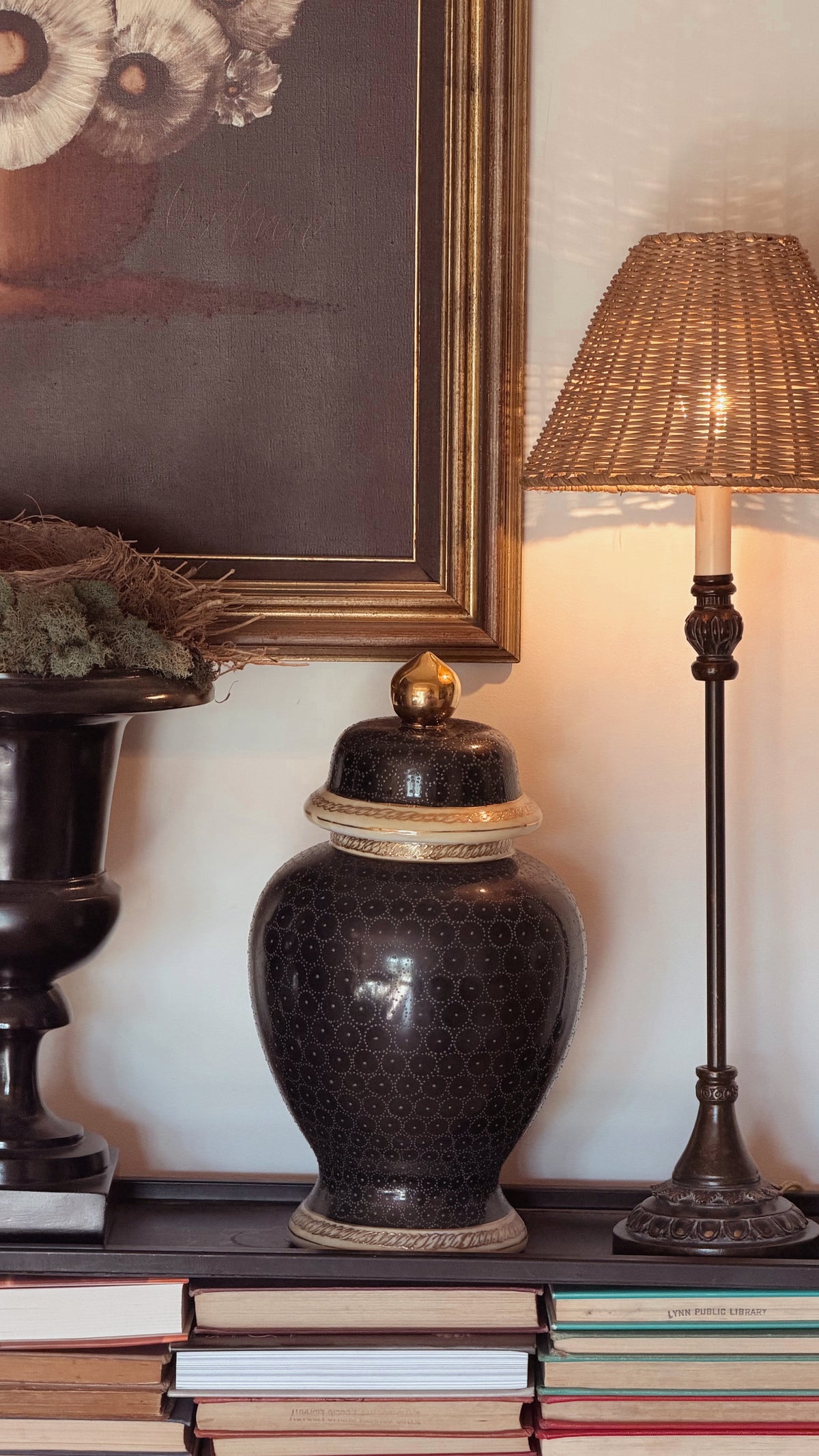 A navy blue patterned ceramic lidded urn with a gold finial sits on a stack of books next to a lit table lamp and a framed painting.