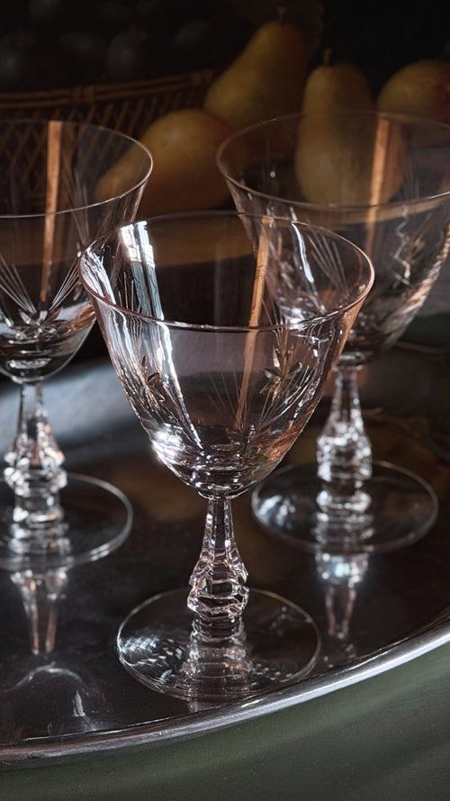 Three clear cut-crystal wine glasses with faceted stems and bases, arranged on a reflective silver tray with softly blurred pears in the background.