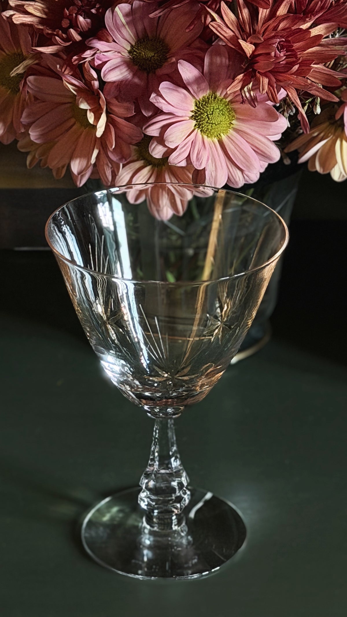 A clear cut-crystal stemmed goblet with a wide flared bowl, shown on a dark surface with pink flowers in the background.