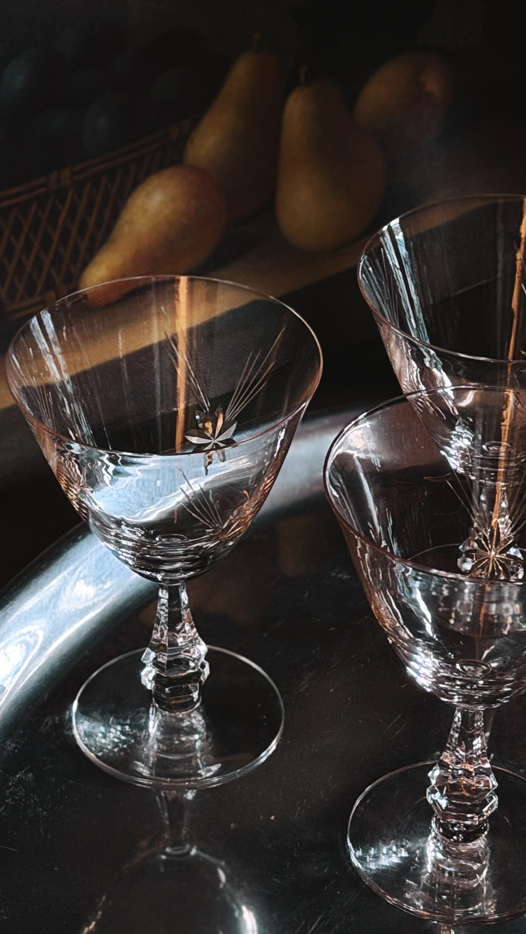 Three clear crystal wine glasses with etched starburst details, arranged on a dark reflective surface with a softly blurred pear in the background.
