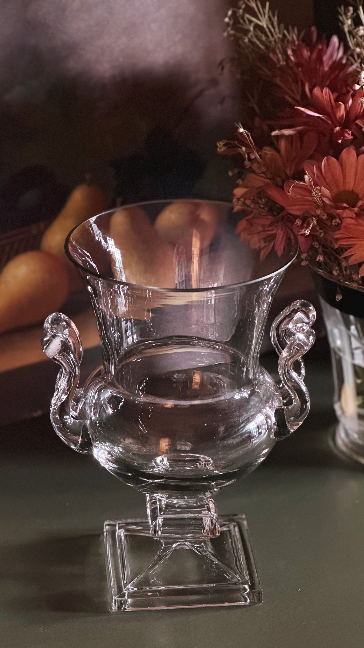 Clear glass urn-shaped pedestal vase with sculpted side handles, shown on a tabletop with a blurred floral and fruit background.