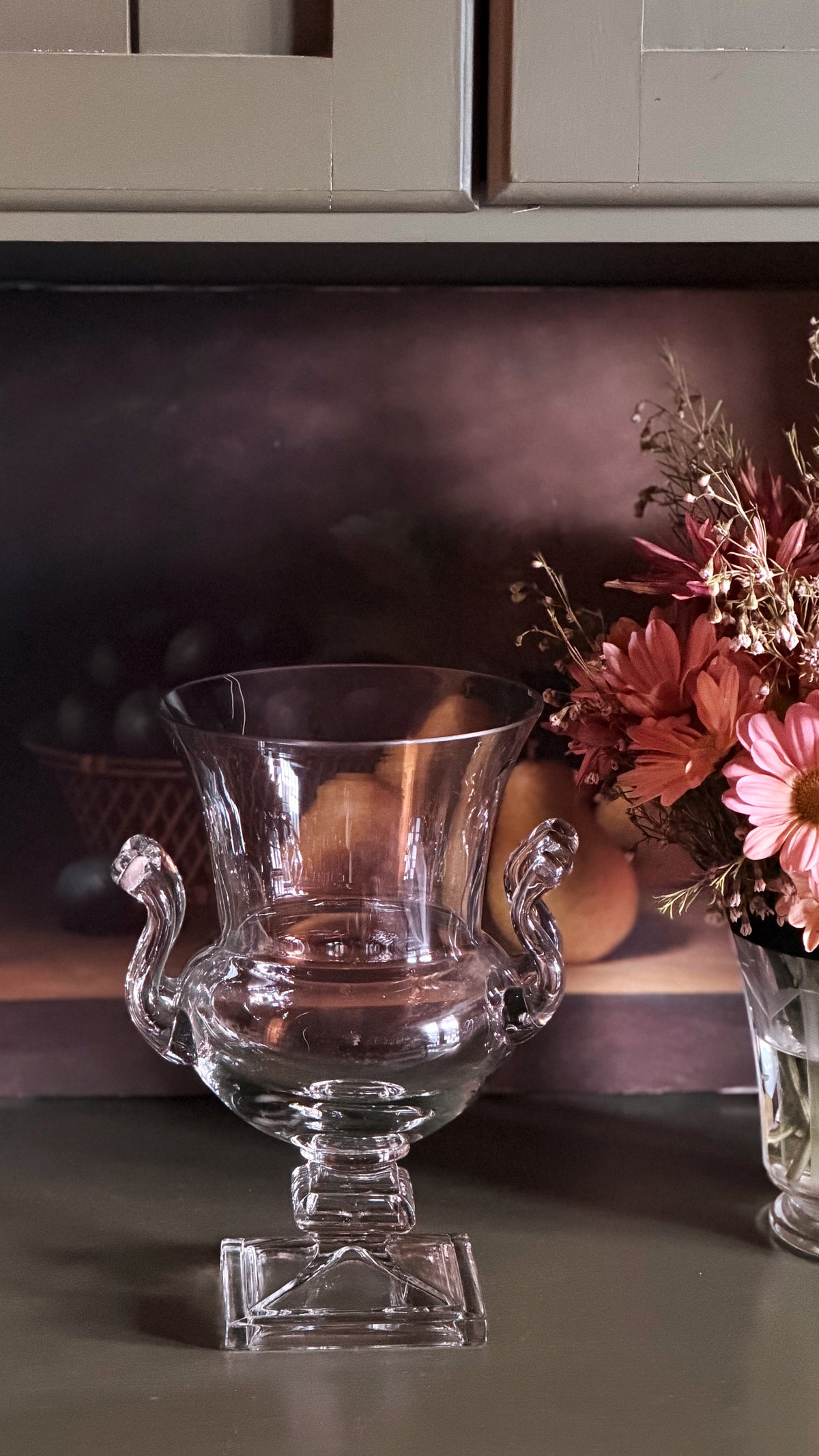 A clear glass urn-shaped vase with two curved handles and a square pedestal base, displayed on a tabletop next to dried pink flowers.