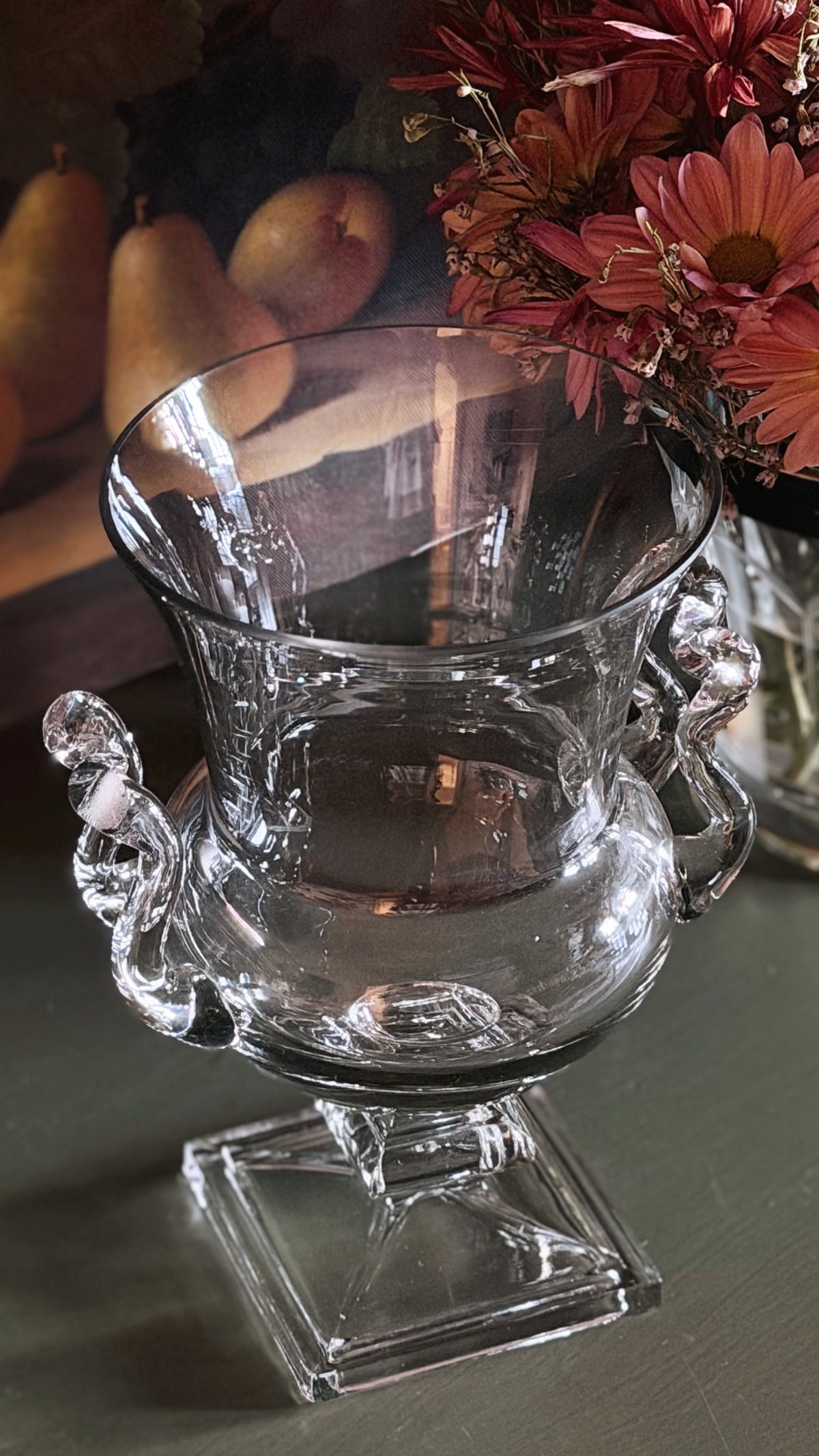 Clear glass urn-style vase with sculpted handles and a square pedestal base, photographed on a tabletop with flowers and fruit in the background.