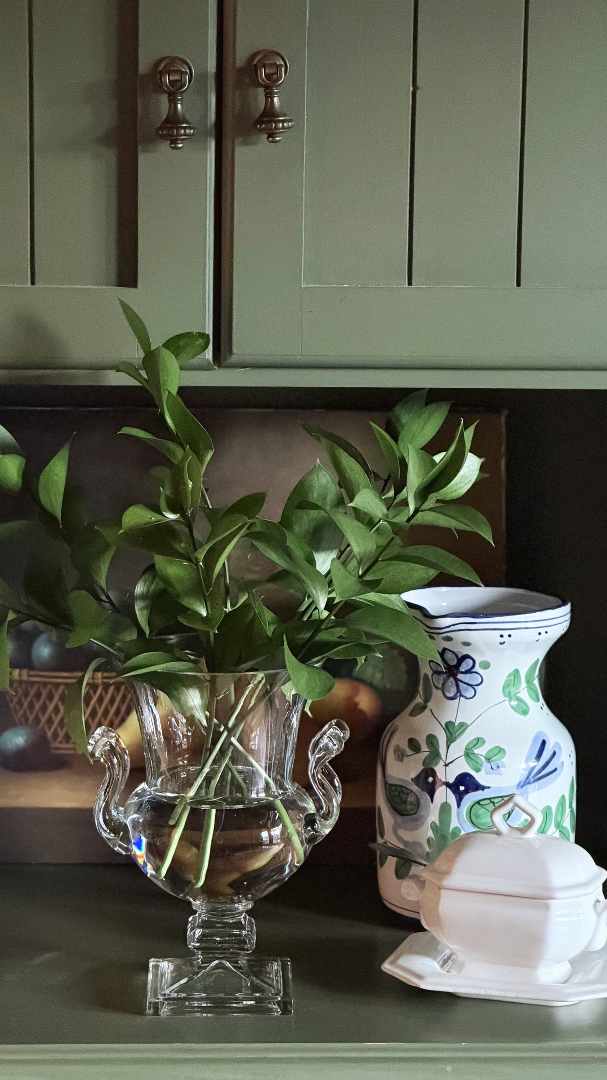A clear glass urn-shaped vase filled with green leafy stems sits on a dark countertop beside a white ceramic pitcher with blue floral and bird motifs, with sage green cabinets in the background.