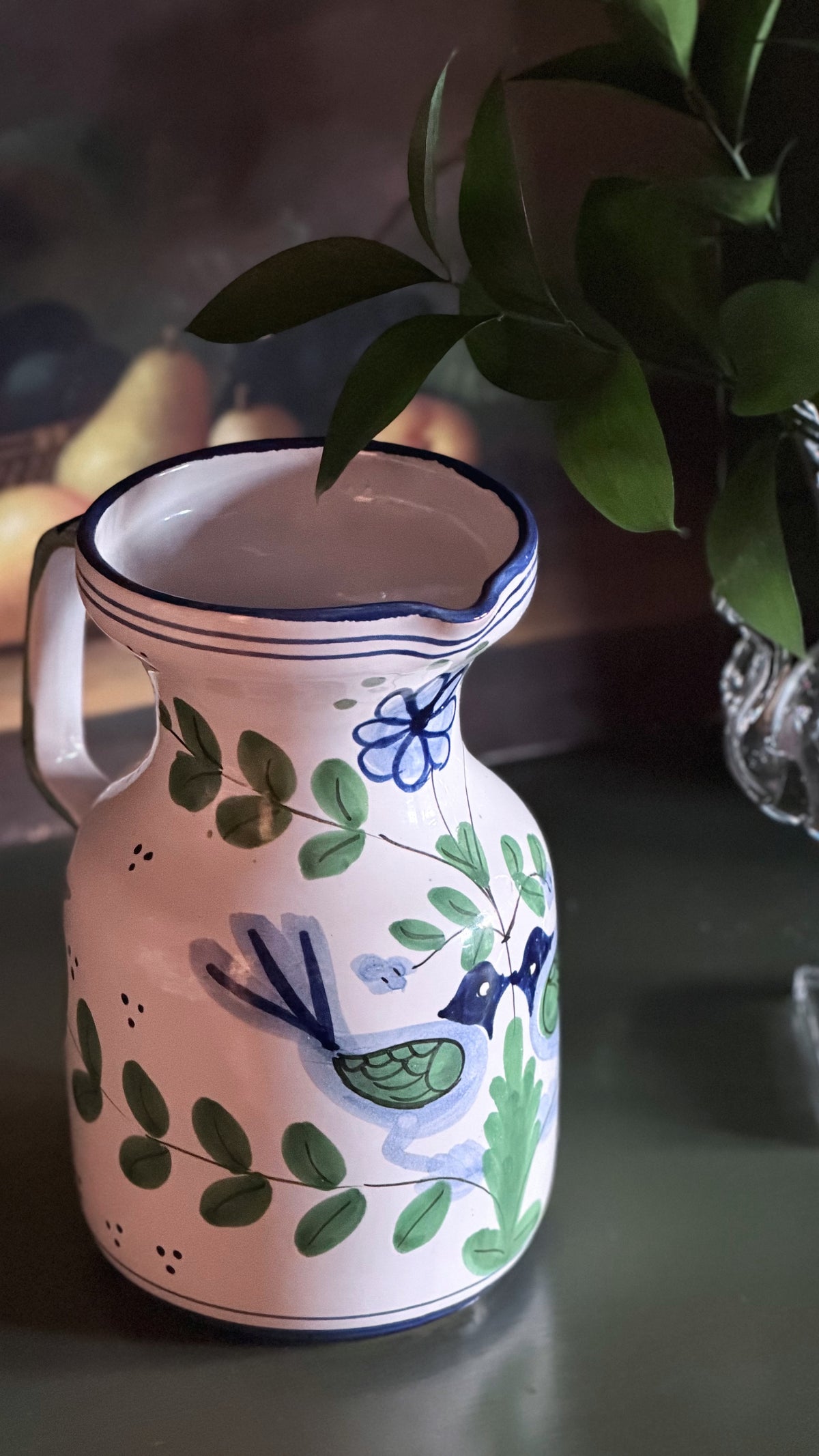 A white ceramic pitcher decorated with green leaves, blue flowers, and a blue-and-green bird, shown on a dark tabletop with a blurred plant in the background.