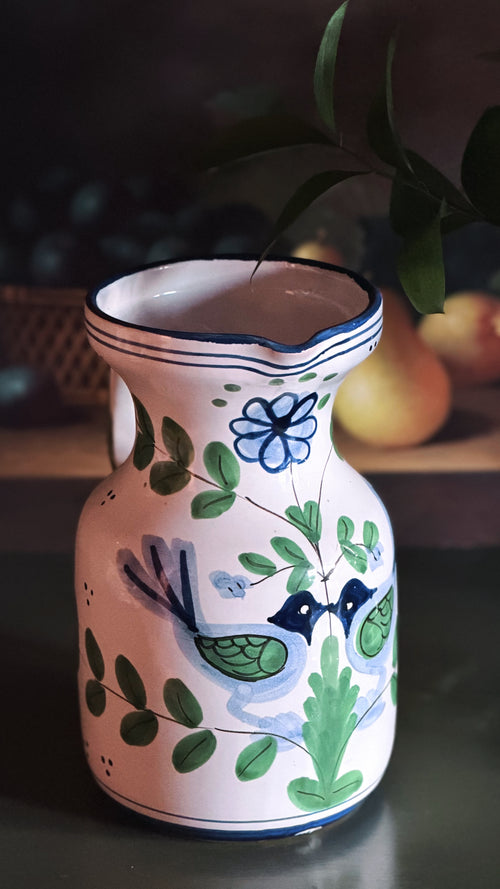 A hand-painted ceramic pitcher with a white base, decorated with green leaves, blue flowers, and two blue birds, set against a dark, softly blurred background with a pear visible.