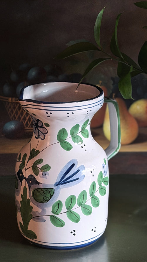 A white ceramic pitcher decorated with hand-painted green leaves, blue birds, and small black dots, set against a dark rustic background with blurred fruit and green leaves.