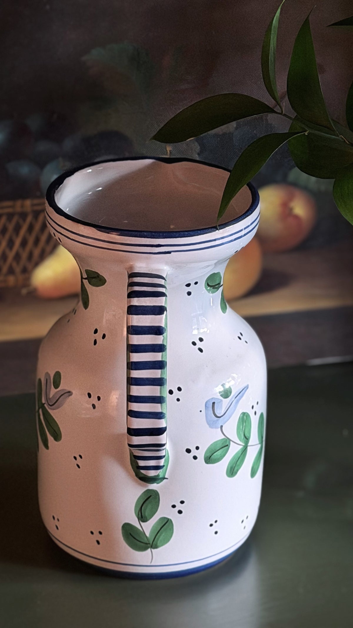 A white ceramic pitcher decorated with green leaves, blue birds, and blue-and-white striped details on the handle and rim, set against a dark, softly blurred background with green leaves in the foreground.