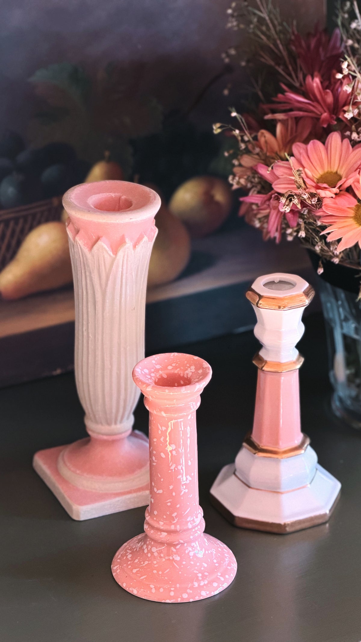 Three decorative pink and white candlesticks arranged on a tabletop, with a blurred floral arrangement and fruit in the background.