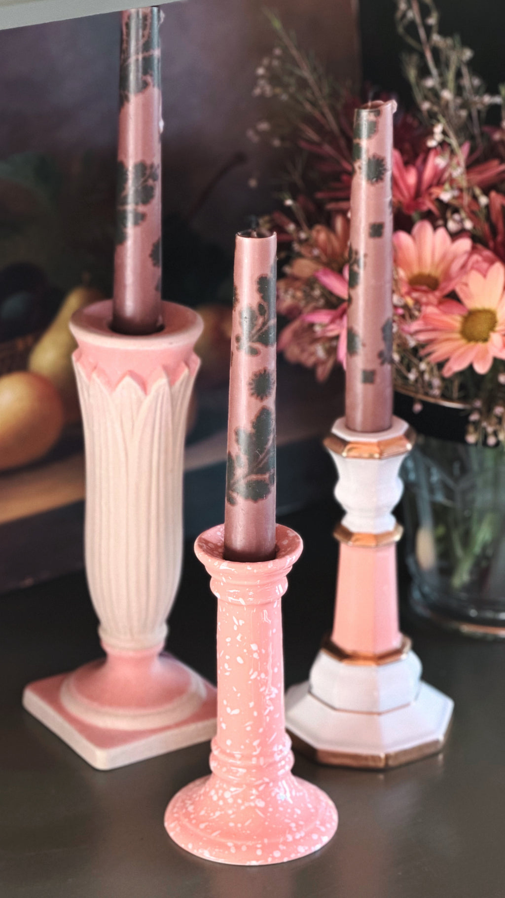 Three pink taper candles with dark floral patterns displayed in decorative pink and white candle holders on a dark tabletop, with flowers in the background.