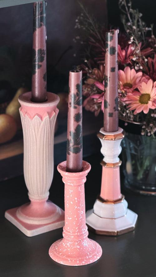 Three decorative pink taper candles with black floral patterns displayed in matching pink and white candlesticks on a dark tabletop, with flowers in the background.