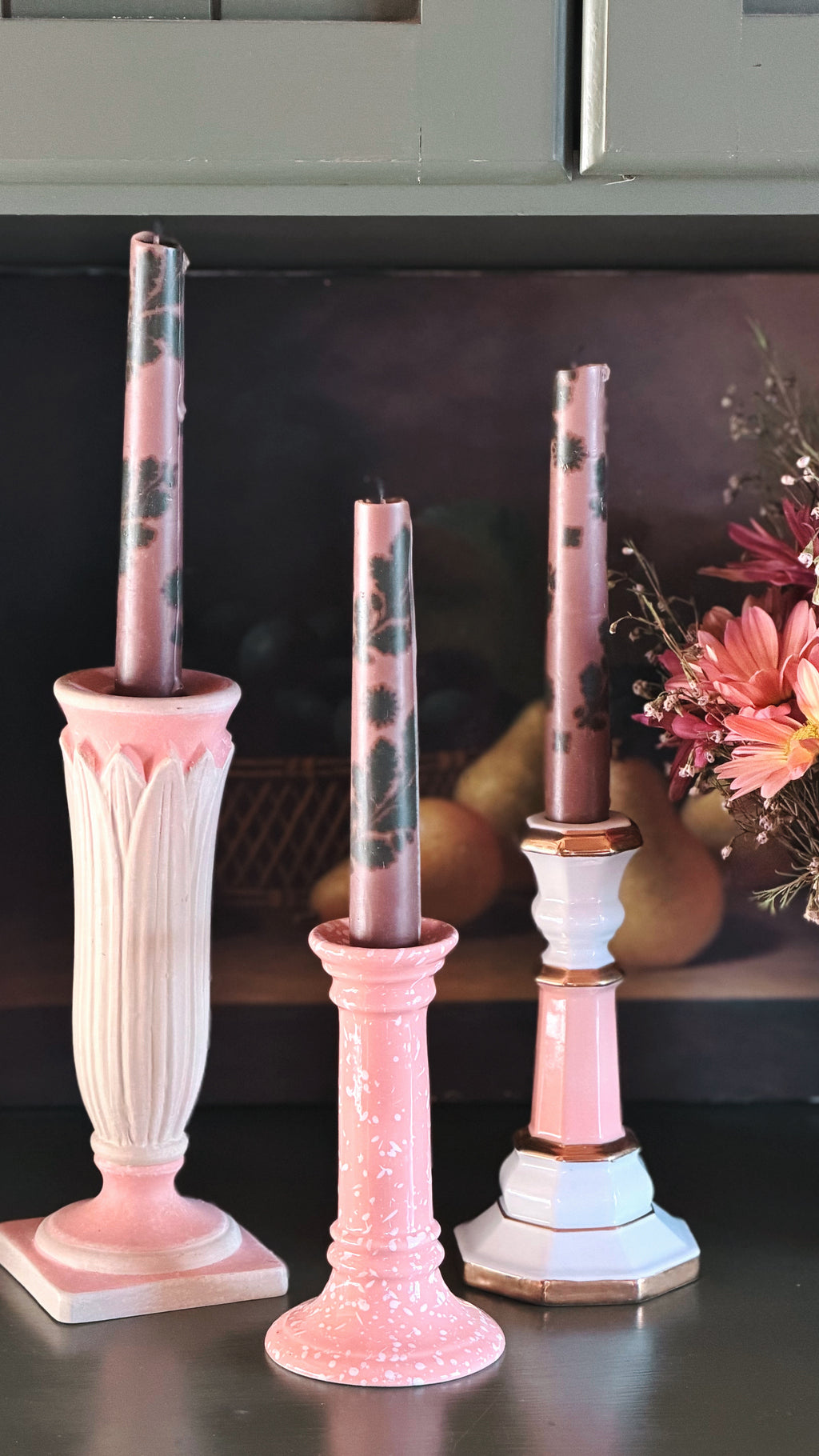 Three pink and white decorative candlesticks with matching patterned taper candles, displayed on a dark tabletop with a blurred kitchen background and flowers at the side.