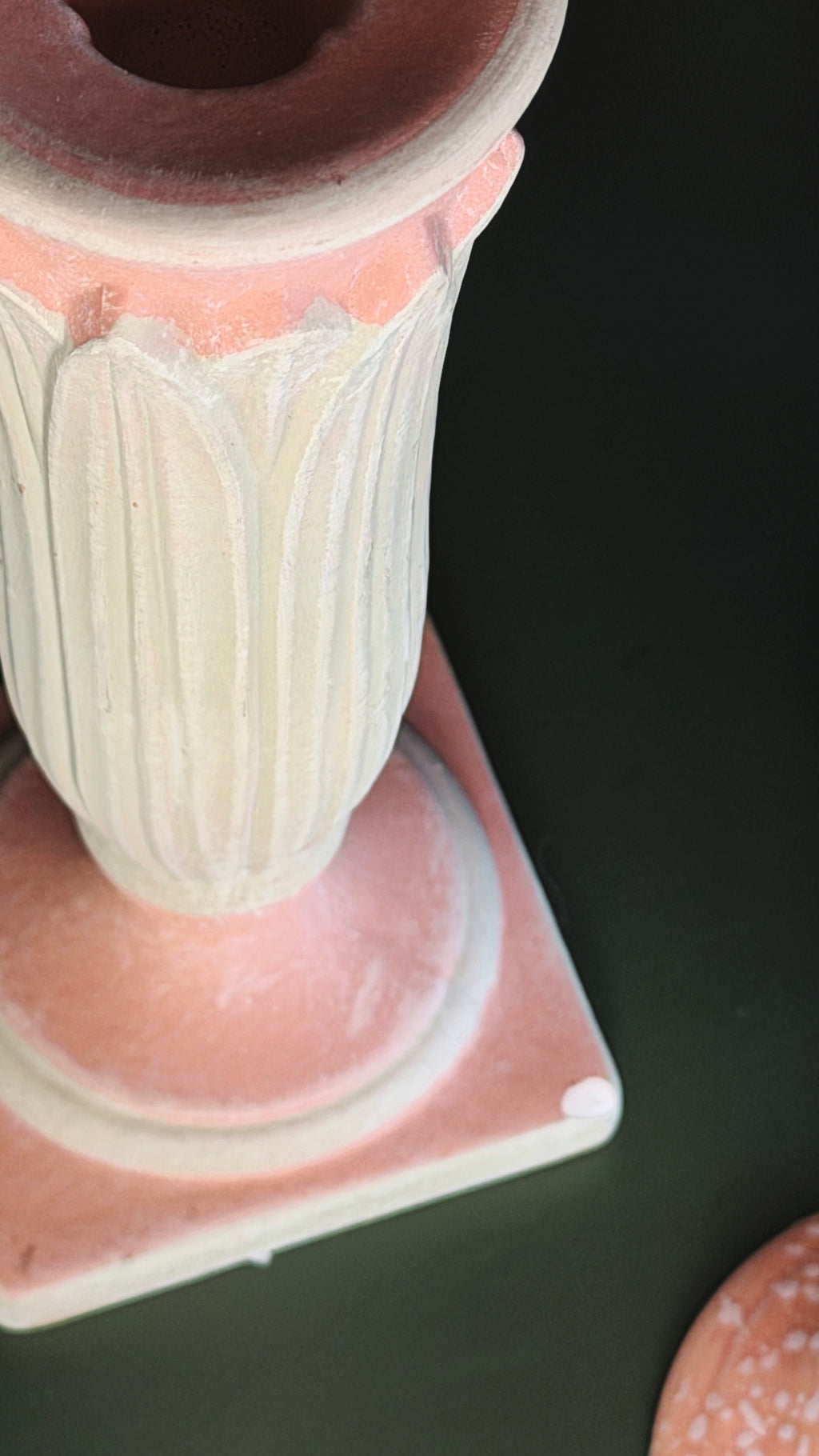 A close-up of a decorative ceramic pedestal vase with a fluted body, a pink base, and a terracotta-colored rim, shown on a dark background.