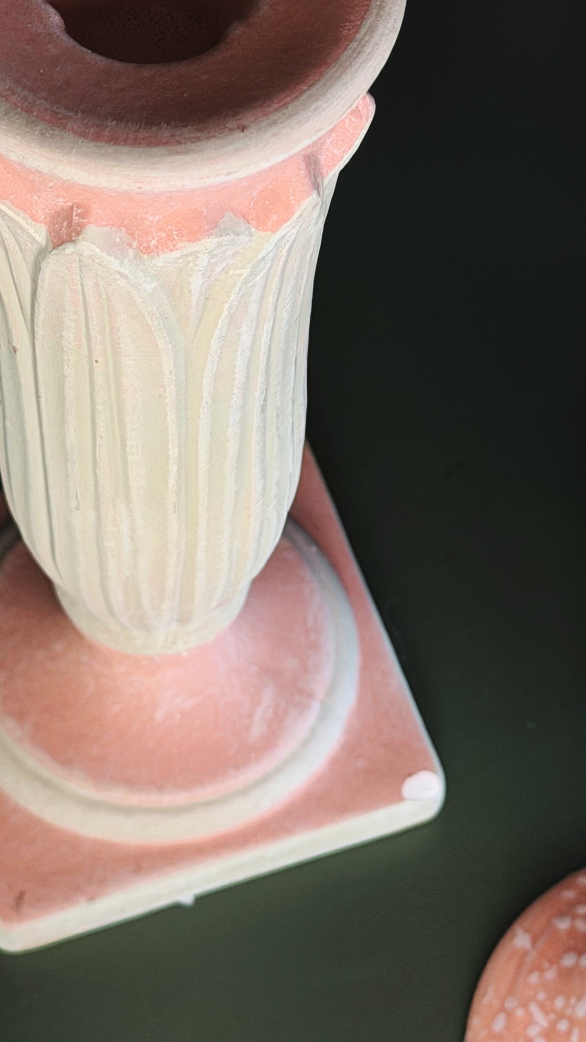 A close-up of a decorative ceramic pedestal vase with a fluted body, a pink base, and a terracotta-colored rim, shown on a dark background.