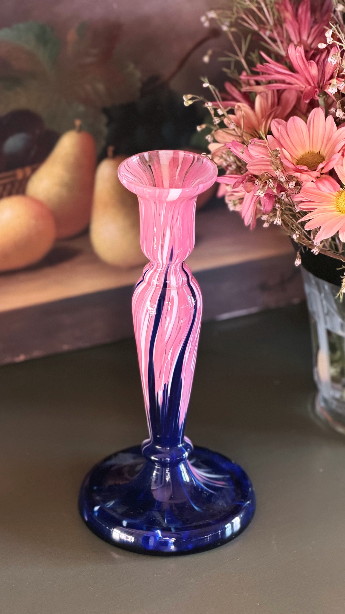 A Pink Cobalt Blue Art Glass Candlestick with a flared top and a round base, set on a tabletop next to a vase of pink flowers.