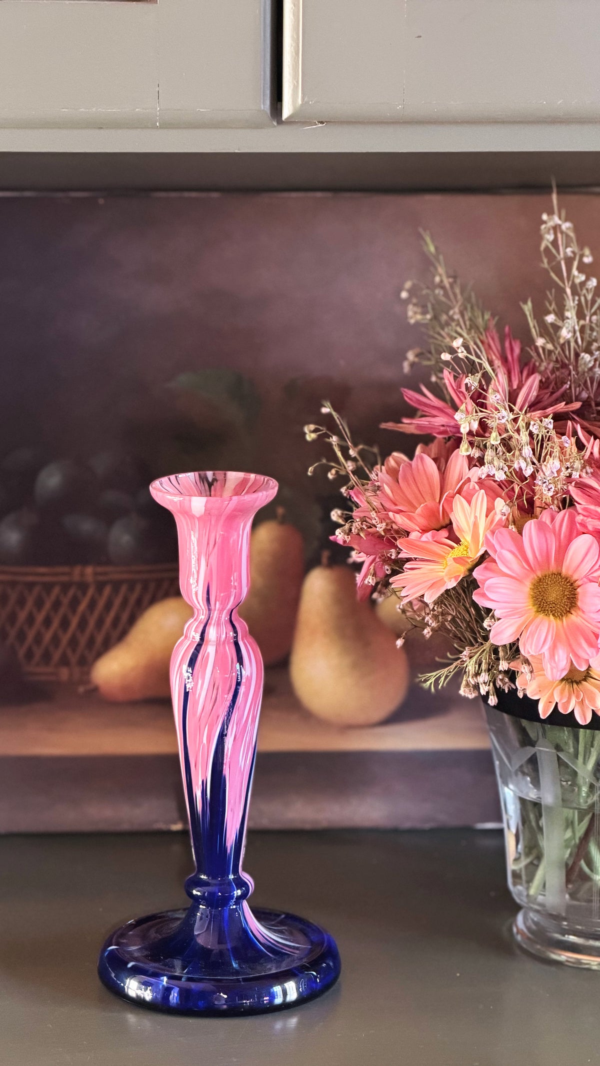 A pink and blue swirled glass candlestick holder with a flared rim, displayed on a tabletop next to a vase of pink flowers against a rustic background.