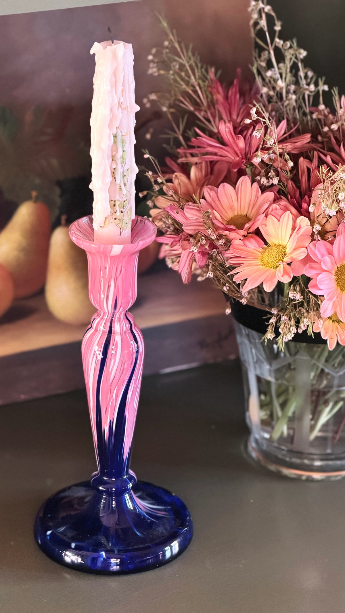 A pink and blue swirled glass candlestick holder with a white candle, styled next to a vase of pink flowers on a tabletop.