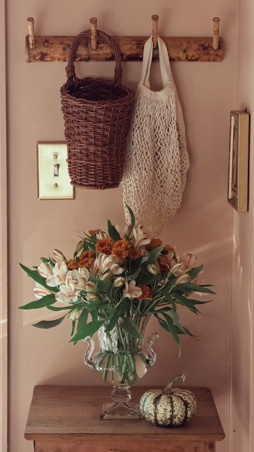 A rustic wooden entryway coat rack mounted on a beige wall, holding a dark brown woven basket and a cream mesh tote bag. Below the rack, a glass vase filled with white and orange flowers sits on a wooden table next to a small decorative pumpkin.