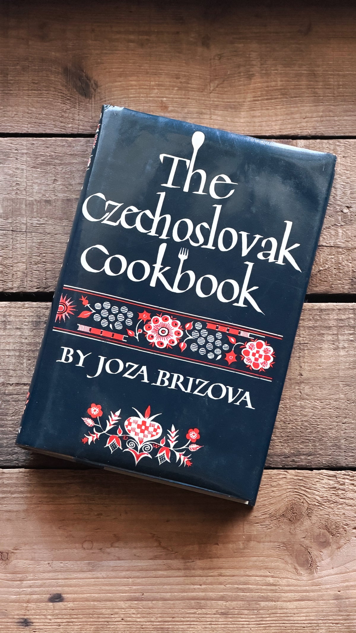 The Czechoslovak Cookbook by Joza Brizova, a dark navy blue hardcover book with white text and red and white floral patterns, resting on a rustic wooden surface.