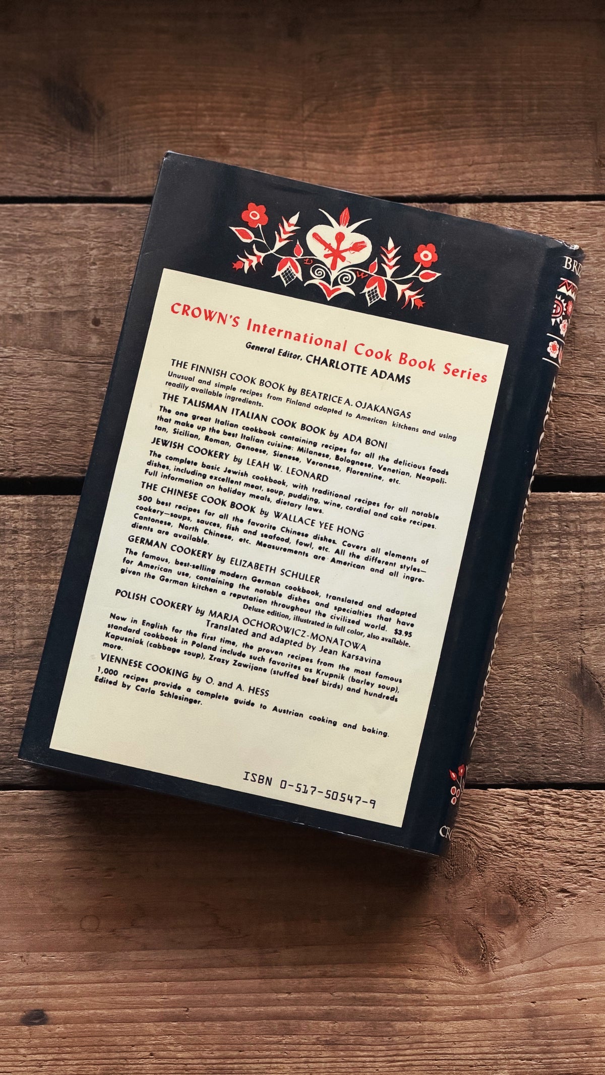 A hardcover cookbook titled 'Crown's International Cook Book Series' by Charlotte Adams, featuring a cream page with black text on the open cover, resting on a dark wooden surface.