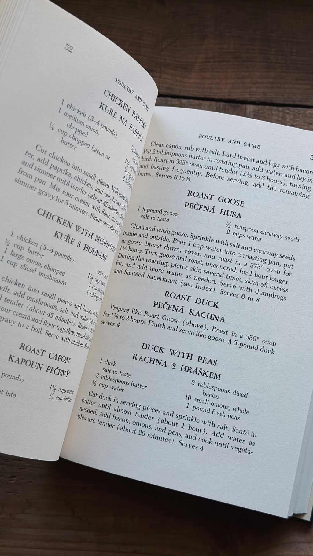 An open cookbook showing printed recipe pages with headings like Chicken with Herbs and Roast Goose, photographed on a dark wooden surface.