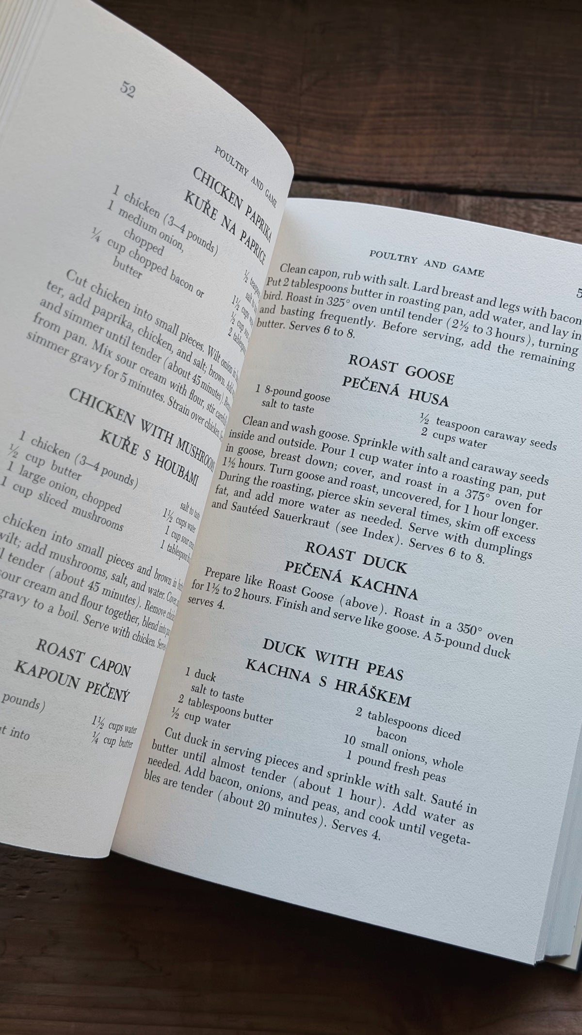 An open cookbook showing printed recipe pages with headings like Chicken with Herbs and Roast Goose, photographed on a dark wooden surface.