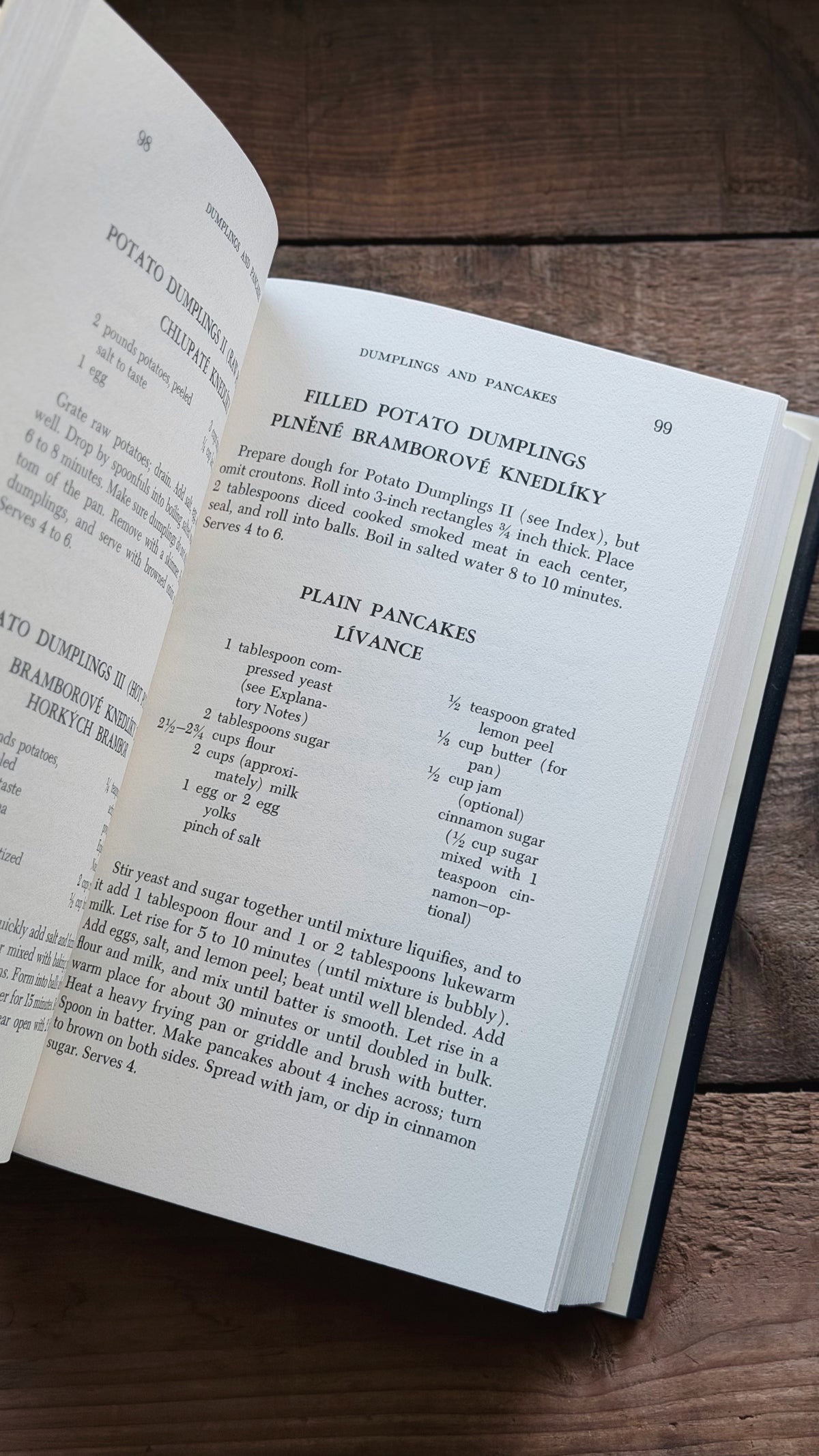 An open cookbook on a dark wooden table, showing printed recipe pages with headings for potato dumplings and pancakes.
