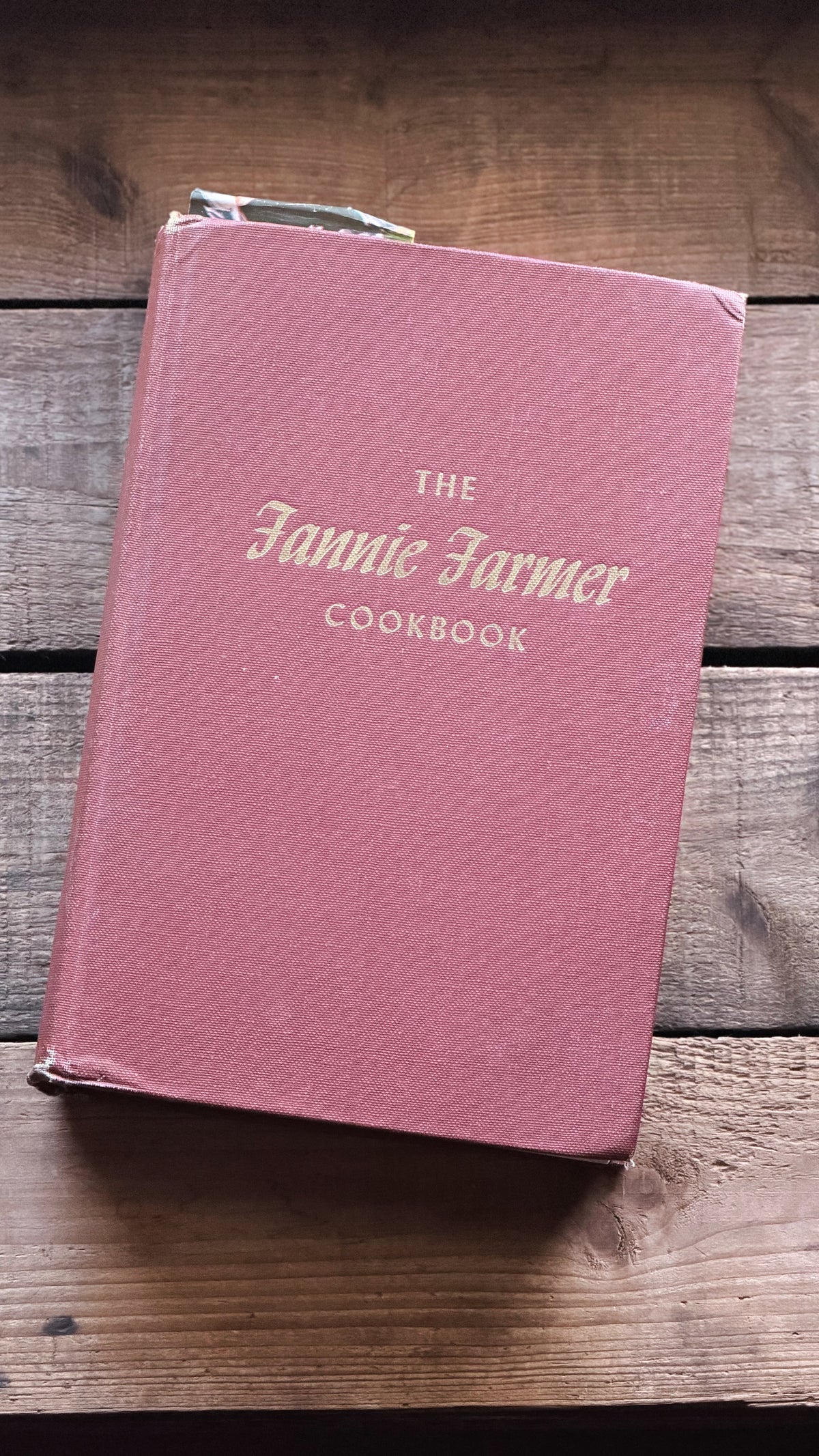 The Fannie Farmer Cookbook in a dusty rose pink hardcover with gold lettering, resting on a rustic wooden surface.