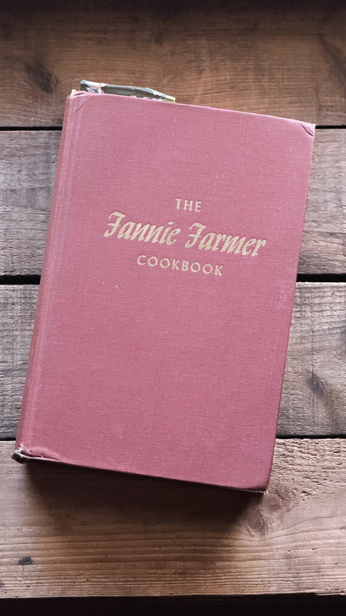The Fannie Farmer Cookbook in a dusty rose pink hardcover with gold lettering, resting on a rustic wooden surface.