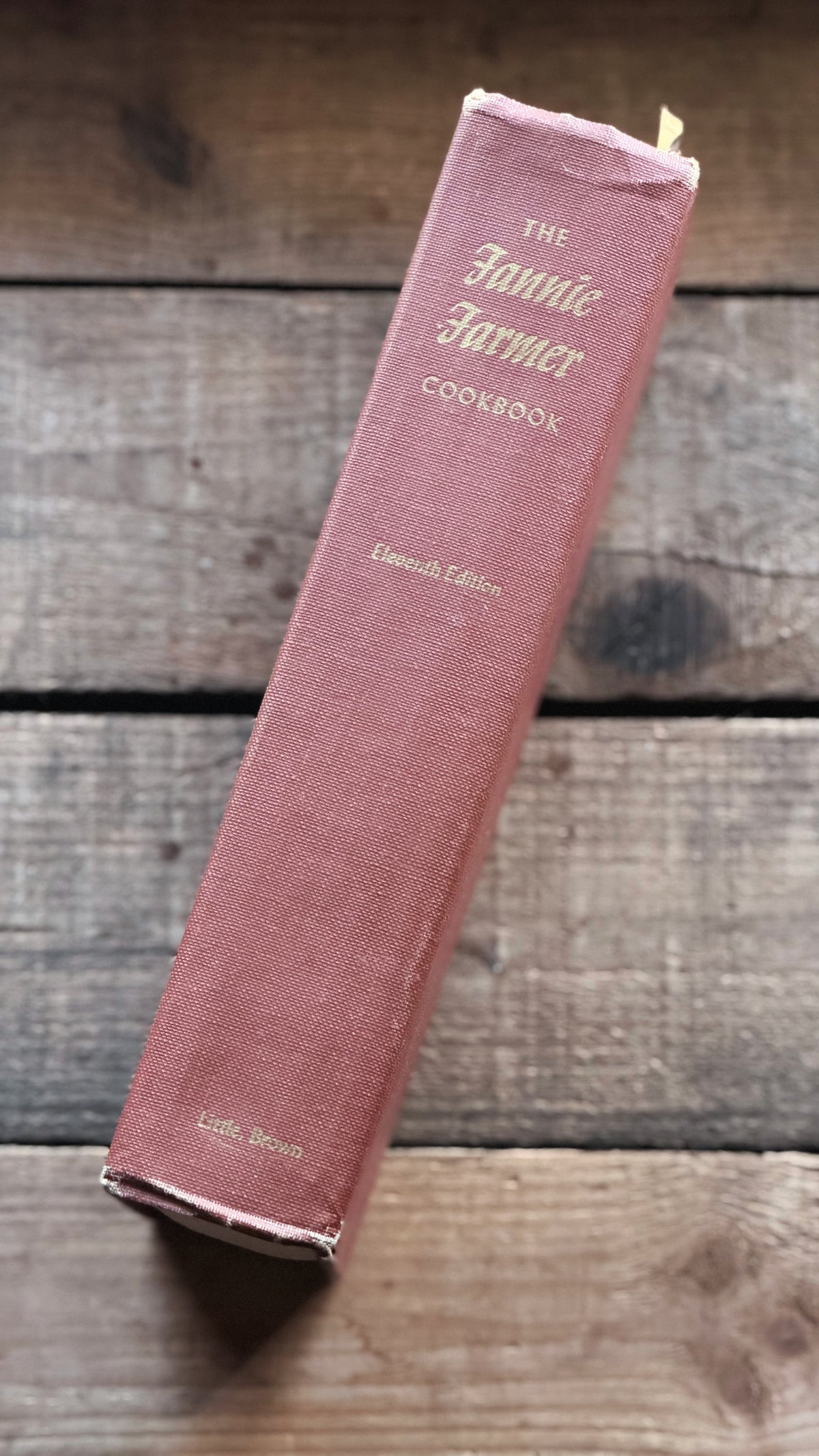 Vintage pink hardcover cookbook titled 'The Fannie Farmer Cookbook' resting diagonally on a rustic wooden plank surface.