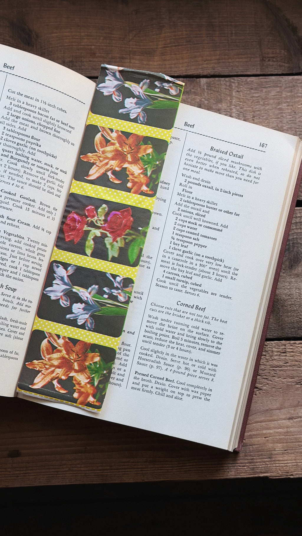 A decorative bookmark with a vertical strip of colorful floral illustrations (orange lilies, red roses, blue flowers) on a yellow patterned background, shown inside an open book on a rustic wooden table.
